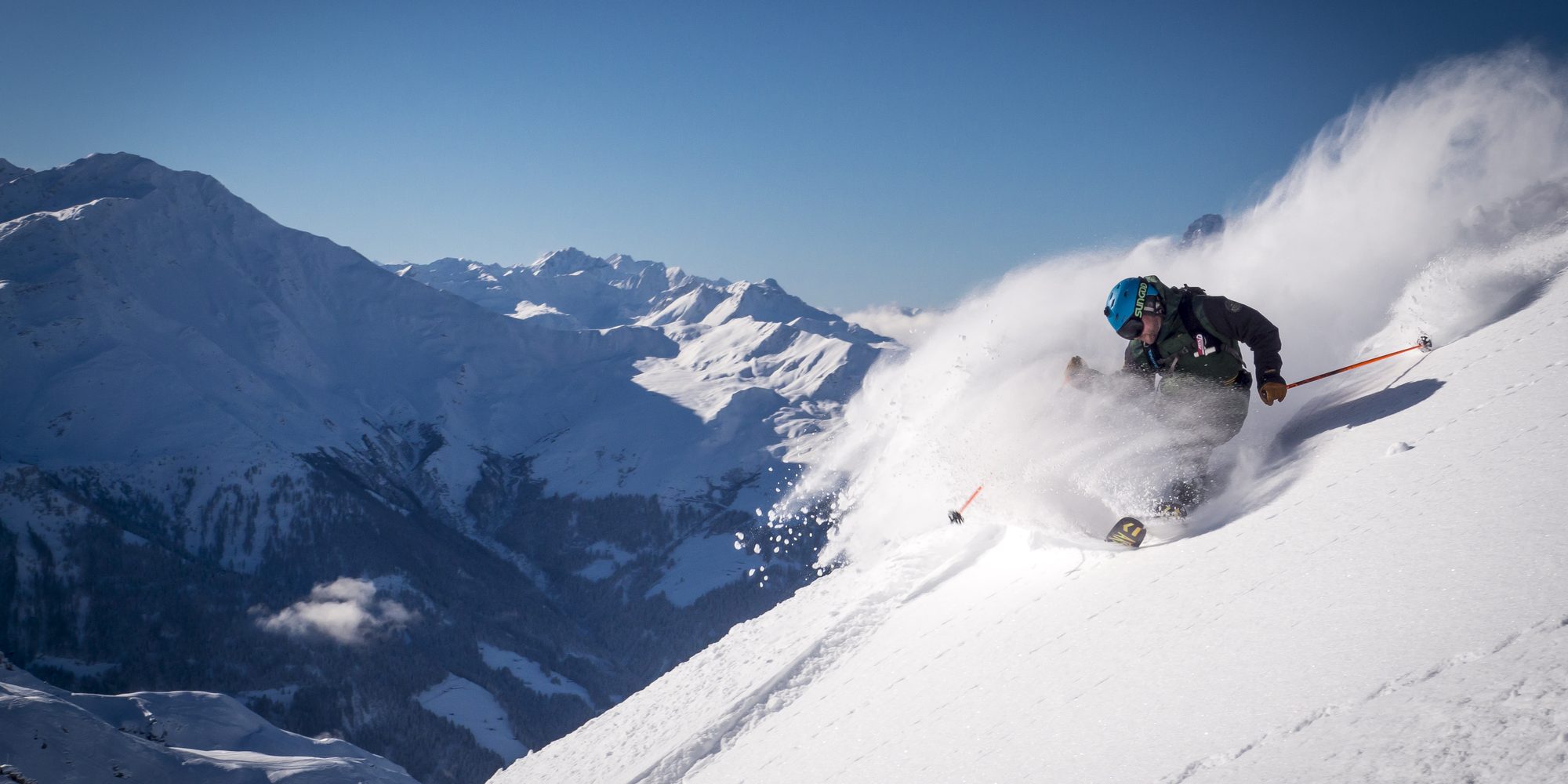 A skier driving through deep powder, spray flying, the Verbier peaks behind