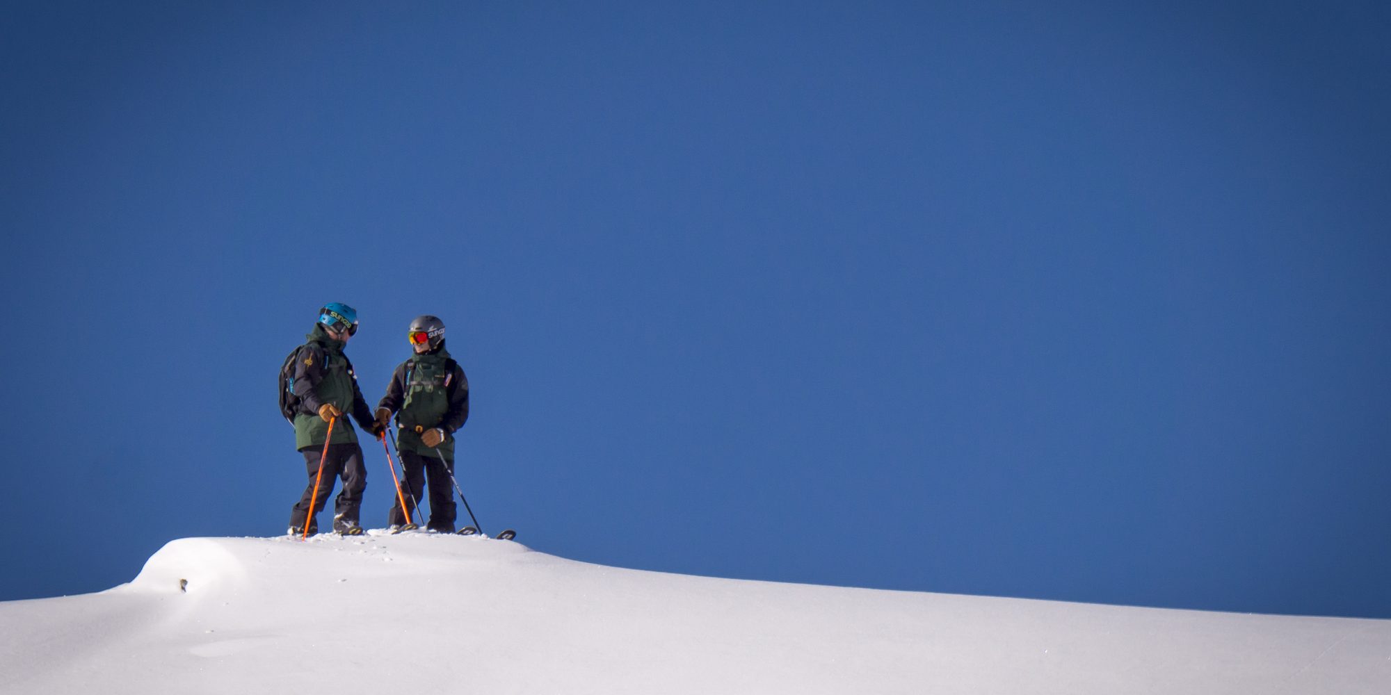 Two ski tourers on a corniced ridge against a deep blue sky